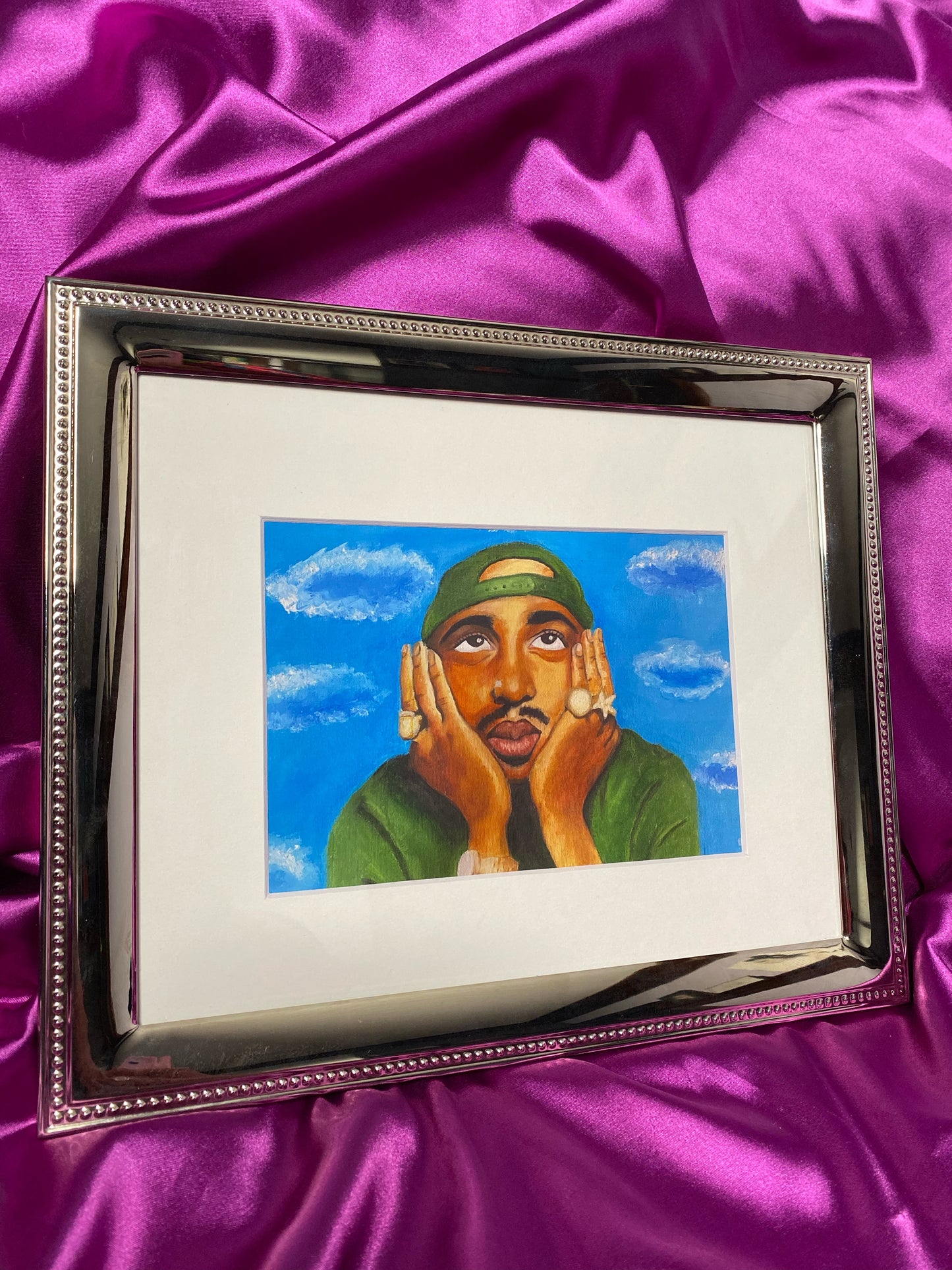 Artwork of Tupac with hands on face against a blue sky with clouds in the background, on a purple fabric background.